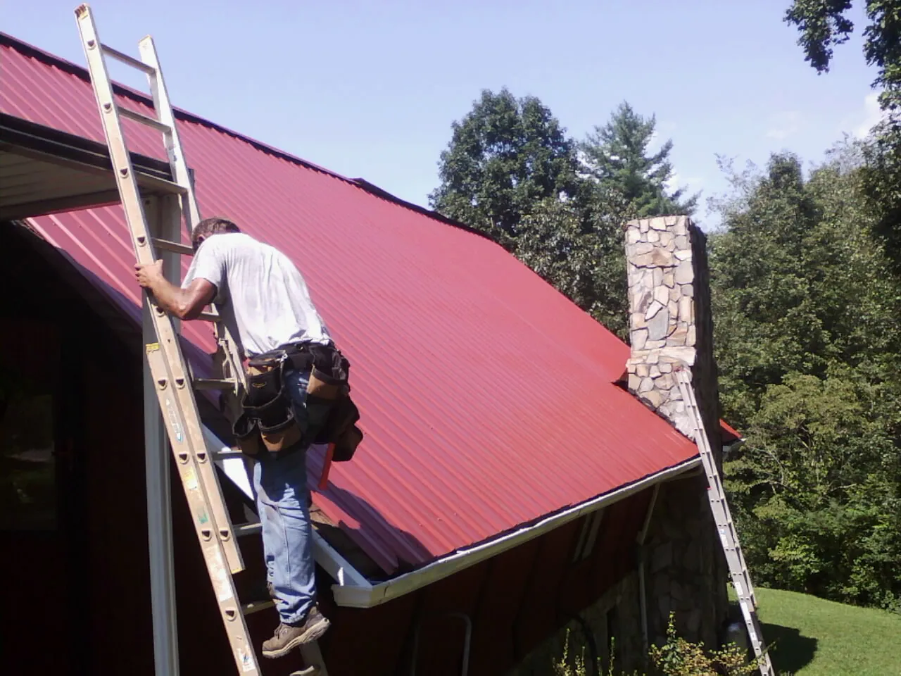 Roofer on a ladder beside a red metal roof and stone chimney — Soffit & Fascia Repair by Roofing Brothers in Reddell